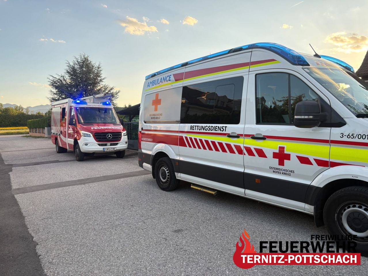 Zwei Krankenwagen sind auf einer Straße geparkt, einer mit blinkenden Lichtern und der andere mit dem Text 'Rettungsdienst'. Der Himmel ist klar mit einigen Wolken.