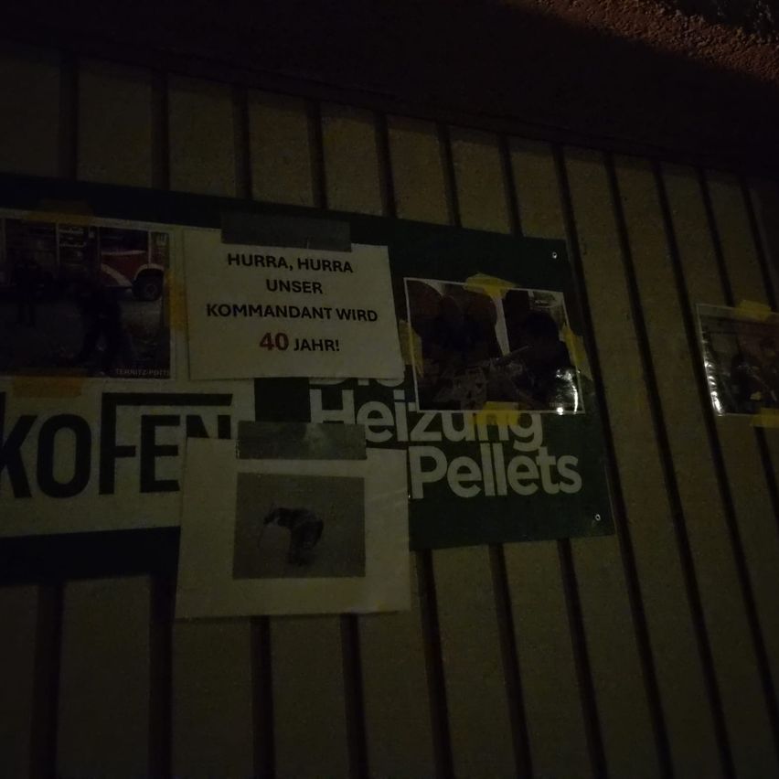 Ein Bulletinbrett mit verschiedenen Plakaten. Eines davon lautet 'Hurra, Hurra, unser Kommandant wird 40 Jahre alt'. Ein anderes lautet 'Heizung Pellets'. In der unteren linken Ecke befindet sich ein Foto einer Person.