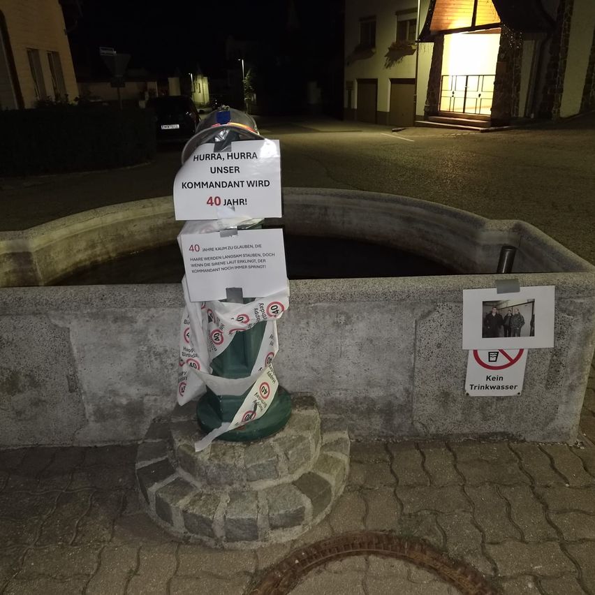 Ein Brunnen mit einem Helm und einem Schild, das 40 Jahre des lokalen Kommandanten feiert. Ein Bild von Menschen ist an der Wand befestigt.