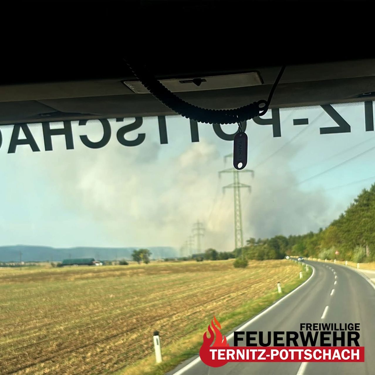 Ein Blick aus einem Fahrzeug zeigt eine Straße mit einem Feuer in der Ferne, begleitet von Rauch und Stromleitungen. Das Bild enthält einen Wasserzeichen, der 'Feuerwehr Ternitz-Pottschach' lautet.