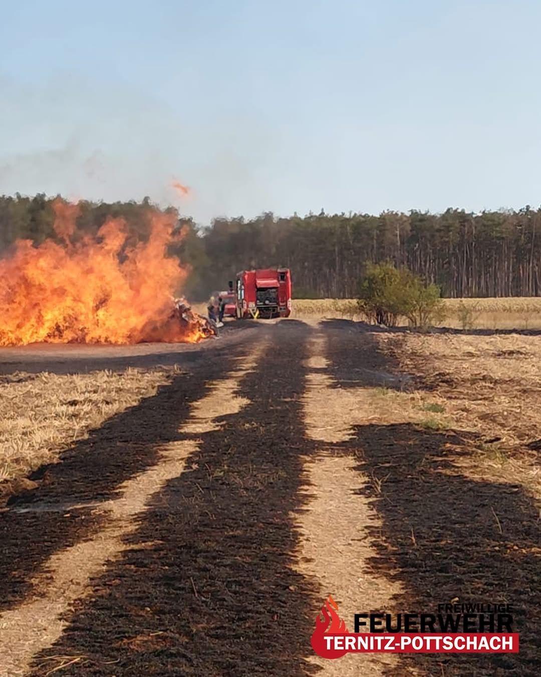 Ein Feuerwehrauto steht auf einem Feldweg, in der Nähe brennt ein großes Feuer. Das Feuer ist von einer verbrannten Fläche umgeben, und das Auto sprüht Wasser.