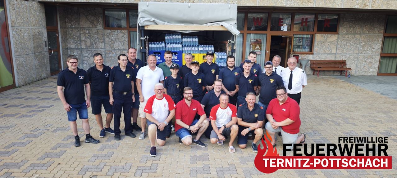 A group of firefighters and volunteers pose for a photo in front of a building with a water storage area.