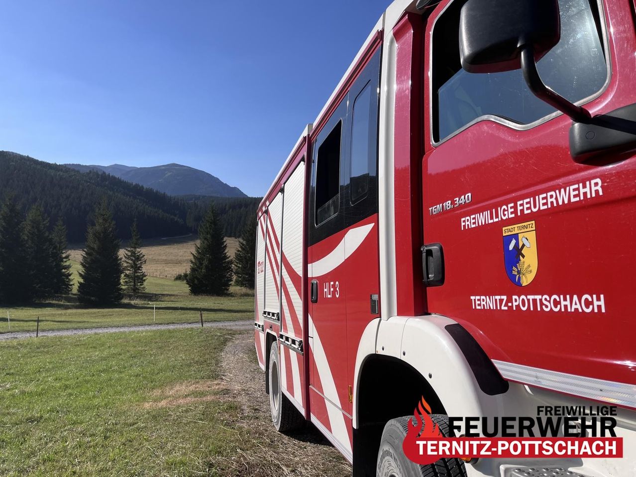 A red fire truck with 'Feuerwehr Ternitz-Potts' written on the side is parked on a dirt road with a grassy field and mountains in the background.