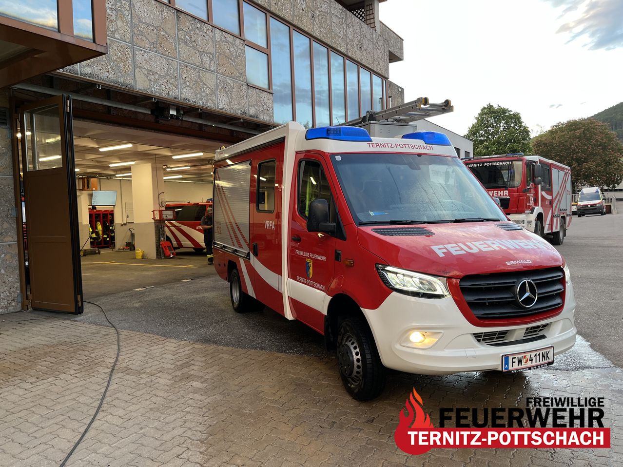A red and white Mercedes-Benz fire truck is parked in front of a modern building with glass walls. The truck has the words 'Feuerwehr' on its side.