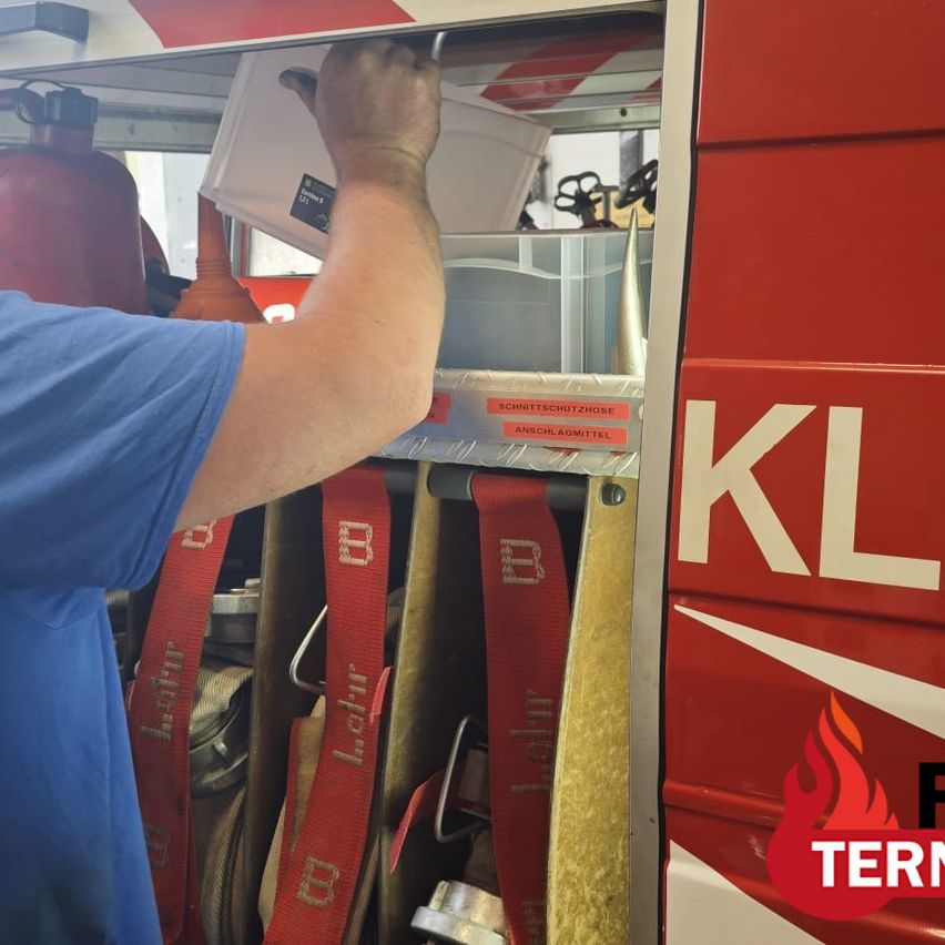 A firefighter is seen inside a fire truck, placing a white container into a compartment labeled 'SCHLAGESCHLAGMOTTEL'. The fire truck is red with the letters 'KL' on its side.