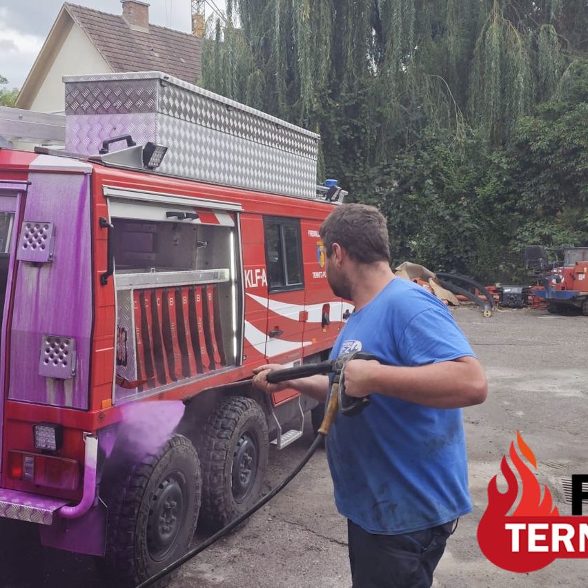 A man in a blue shirt is cleaning a red fire truck with a hose. The truck has a purple accent and is parked in a lot with trees and a house in the background.