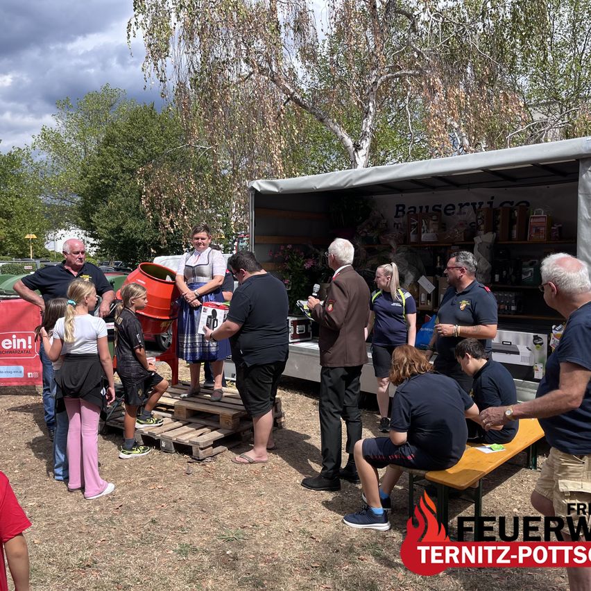 A group of people are gathered around a food truck, with some standing on wooden pallets and others sitting on a bench. The food truck is parked under a tree.