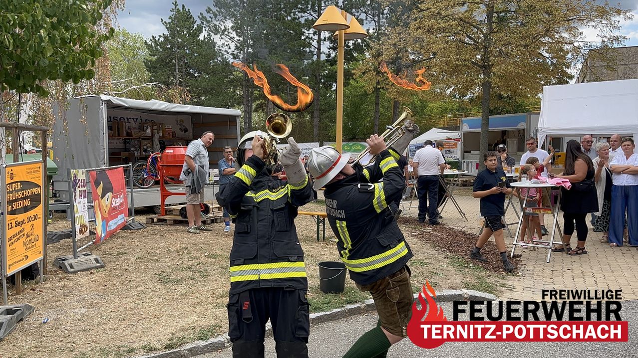 Two firefighters playing trumpets with fire coming out of the instruments, surrounded by a crowd and outdoor setting.
