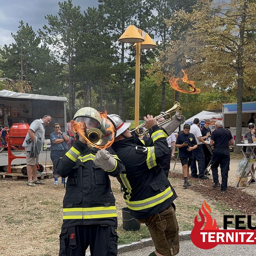 Two firefighters in full gear play trumpets with flames coming out of the instruments. Spectators watch in the background.