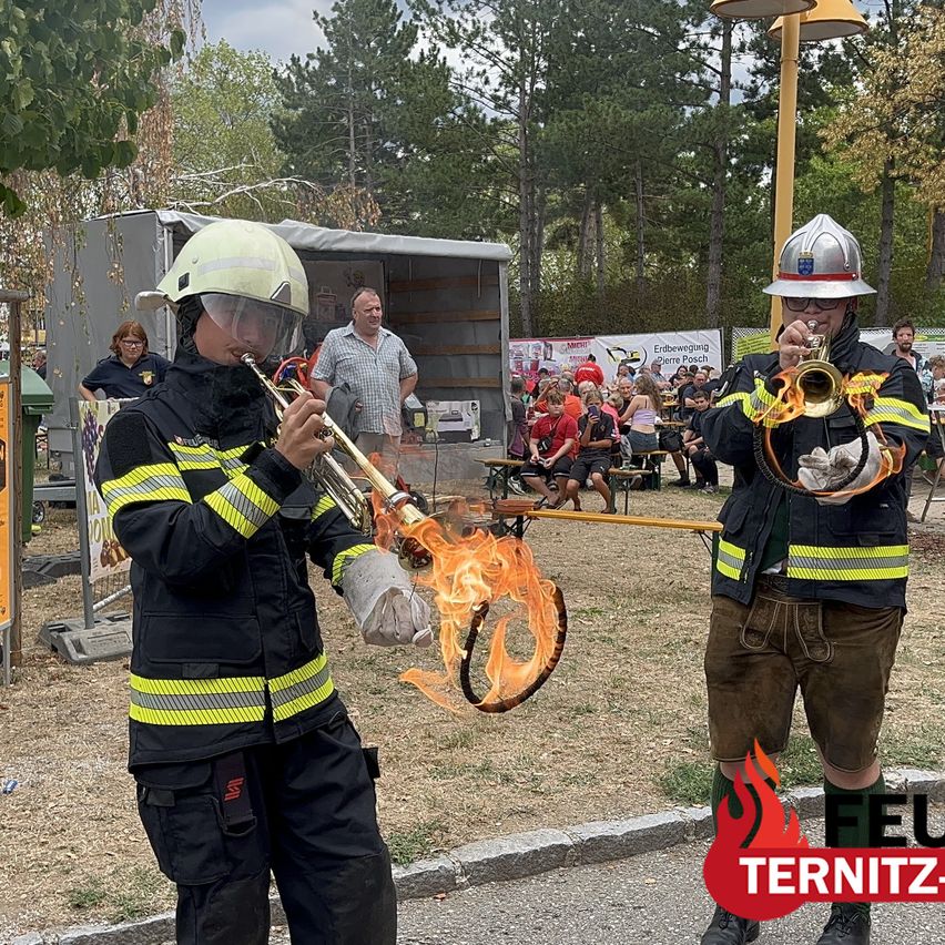 Two firefighters in full gear play trumpets with flames coming out of the instruments, standing on a road with a crowd in the background.