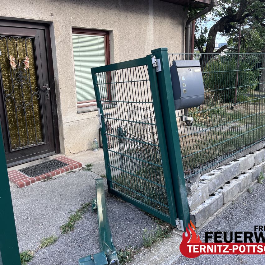 A green metal gate is open in front of a house with a mailbox attached to the fence. A brick walkway leads to the entrance.