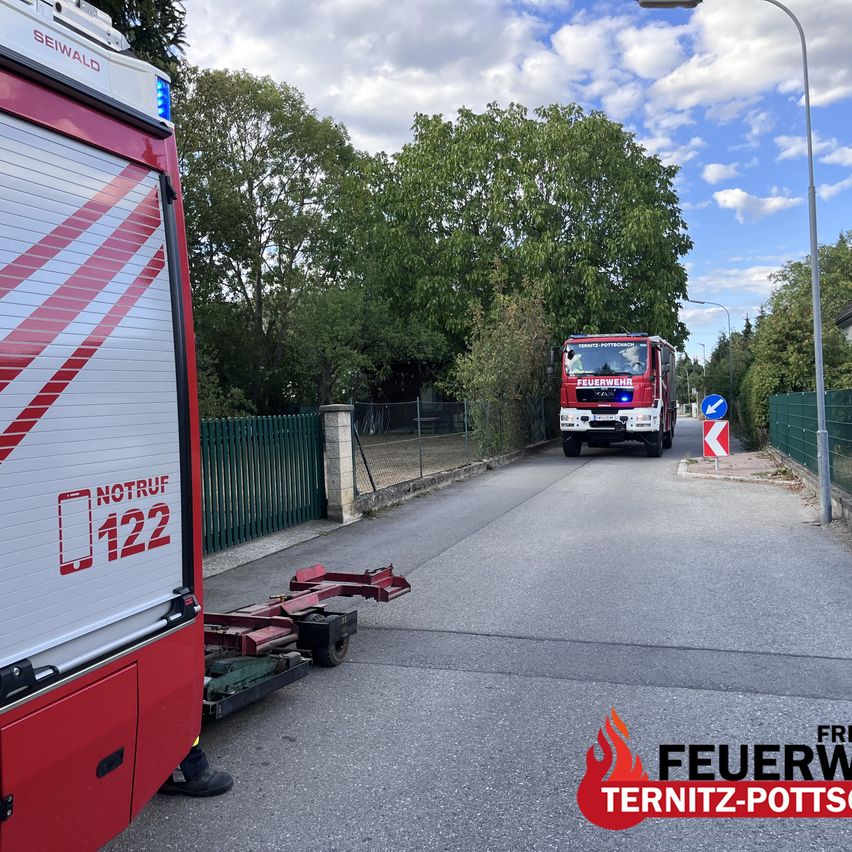 A red fire truck with the number 122 on the side is driving down a street with a green fence and trees on the side.
