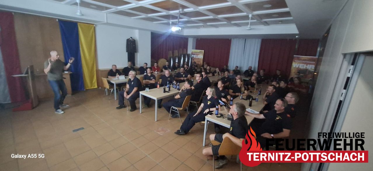 A group of firefighters in uniform are sitting around tables in a room, with a sign reading 'FEUER' in the background. They are engaged in a meeting or training session.