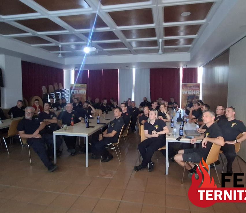 A group of firefighters in uniform are gathered in a room, sitting around tables with bottles and papers. A projector is on the ceiling, and banners are on the wall.