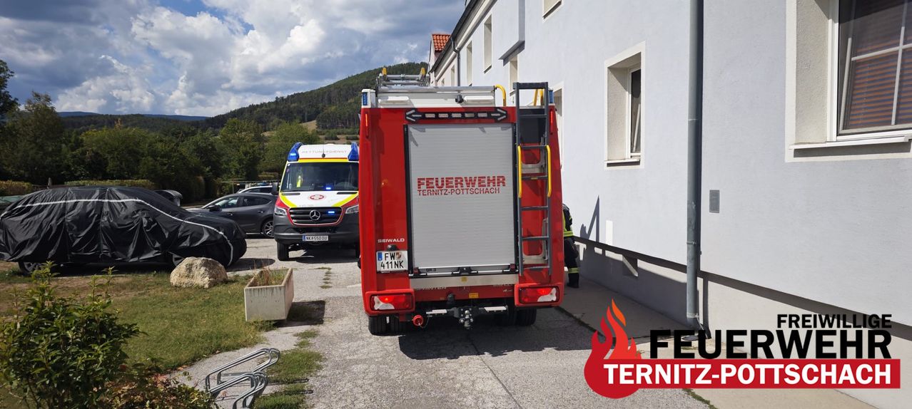 A red fire truck with the word 'Feuerwehr' on the back is parked in front of a building. A person in a yellow vest is standing near the fire truck.