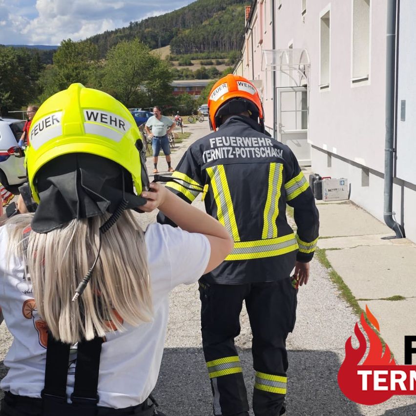 A young girl wearing a yellow firefighter helmet is being assisted by a firefighter in a yellow and black uniform. They are walking on a sidewalk next to a building.