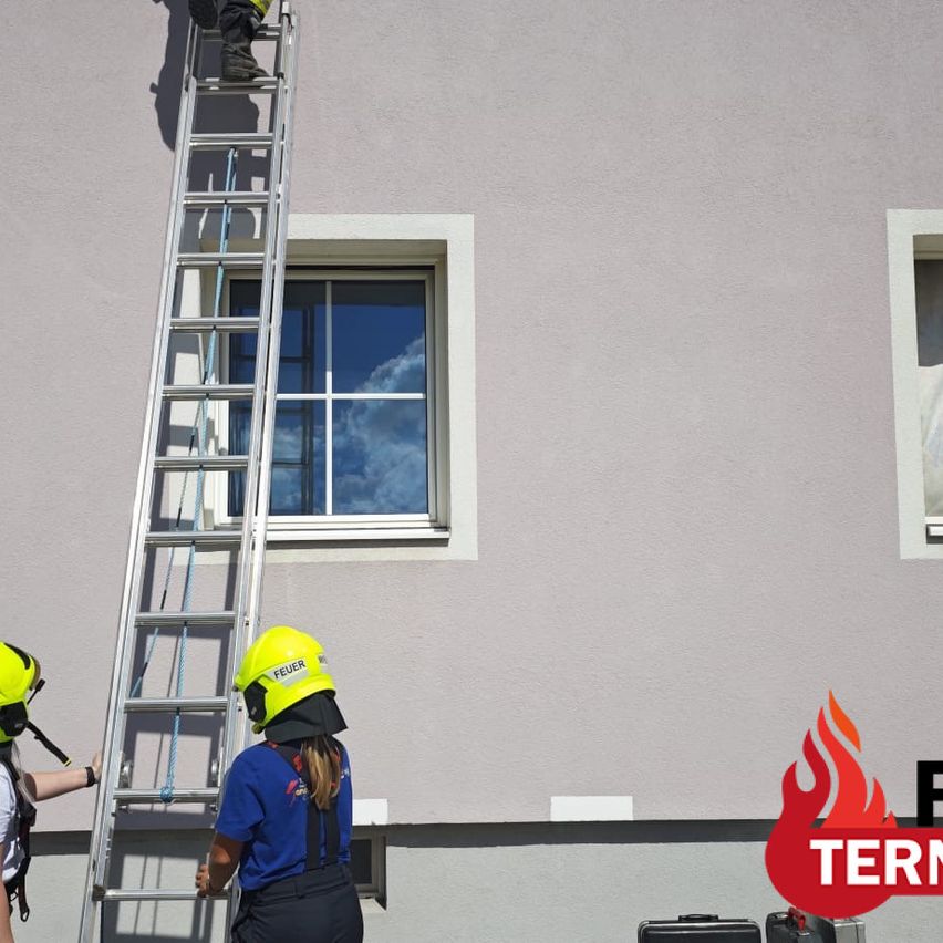 Two firefighters in yellow helmets are climbing a ladder against a building with a window reflecting the sky.