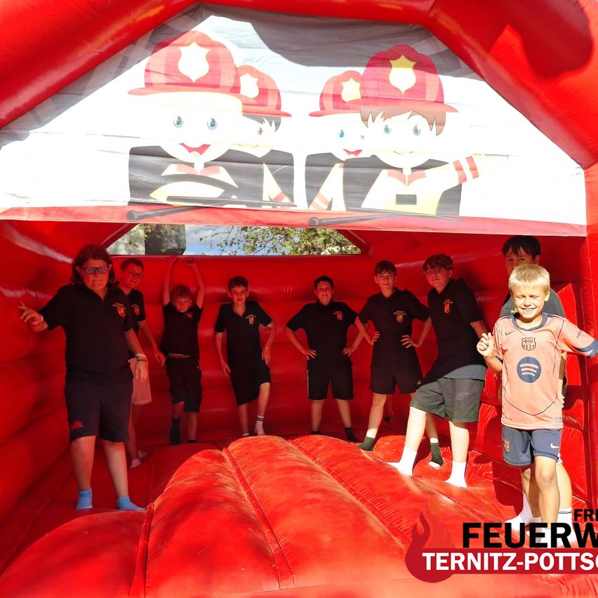 A group of children dressed as firefighters are playing in a red inflatable bounce house. The inflatable is decorated with firefighter illustrations.