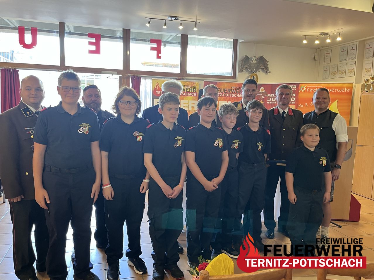 A group of young people in uniform, possibly firefighters, stand together in a room with banners and a fire extinguisher in the background.