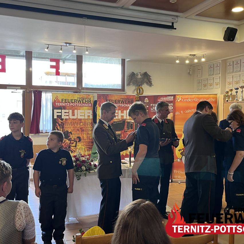 A group of people, including children, are standing in a room with banners and a table with flowers. A man in uniform is giving an award to a boy.