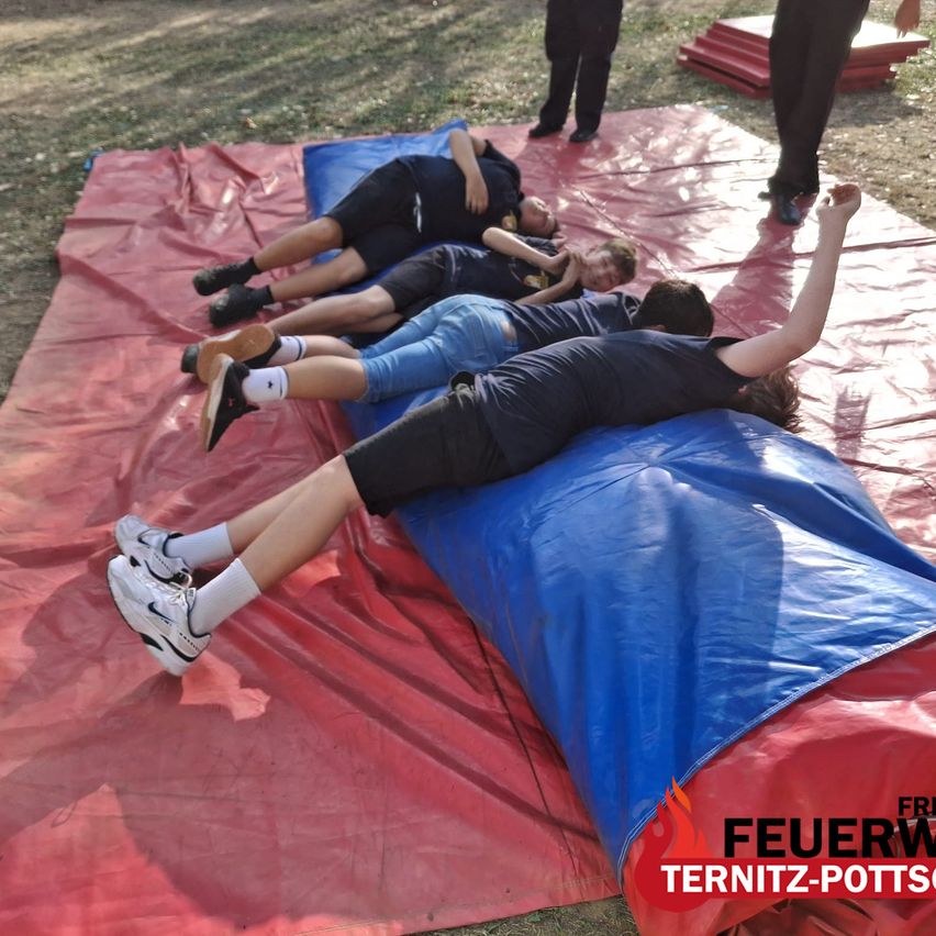 Four individuals lie on blue tarps on a red surface, possibly participating in a training exercise. Two people in the background are standing and observing.