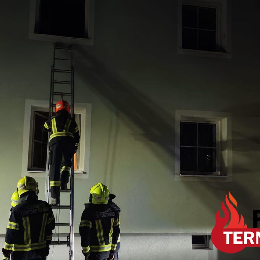 Firefighters in yellow and black uniforms are climbing a ladder to a window on a building at night.