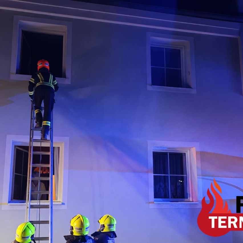 A firefighter climbs a ladder to a window on a building, while others stand below. The scene is illuminated by blue lights.