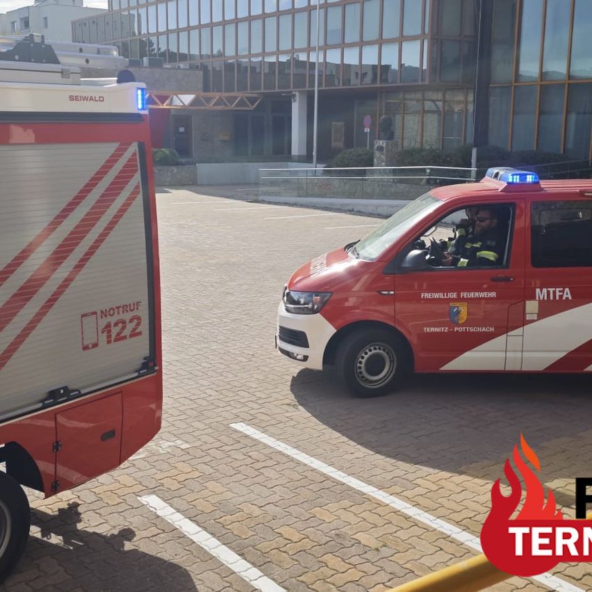 A red fire truck with 'NOTRUF 122' on the side is parked in front of a modern building. A firefighter is sitting inside the vehicle.