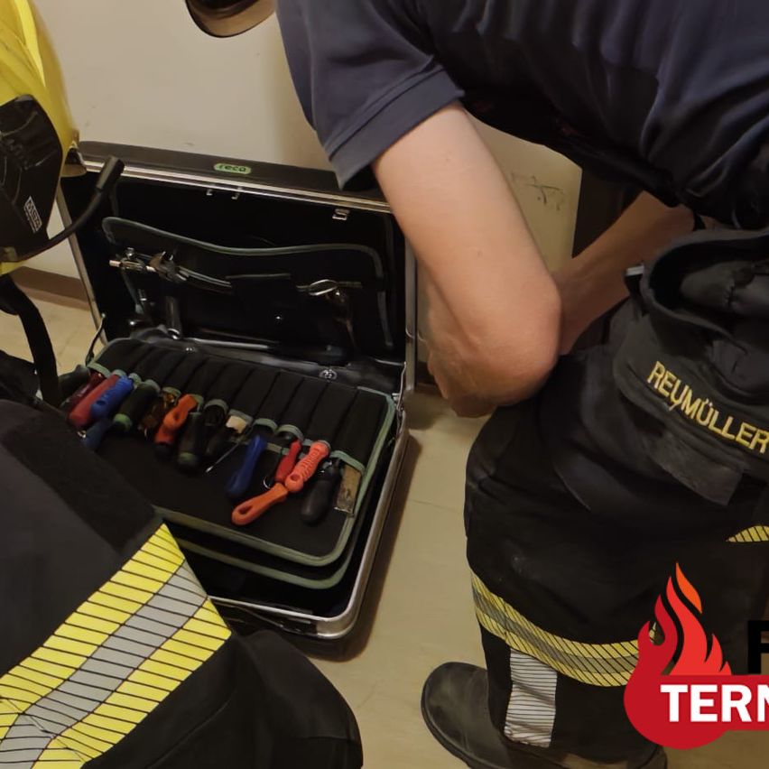 A firefighter with a tool kit open on the floor, another firefighter in yellow uniform beside him.