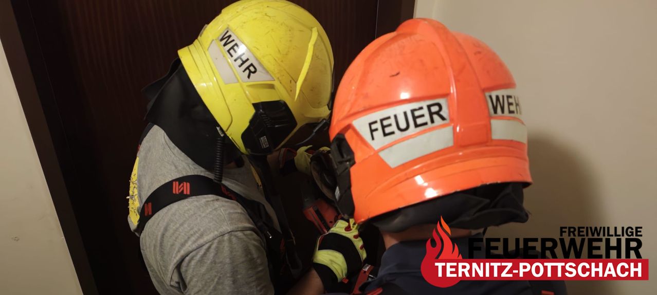 Two firefighters in protective gear, one with a yellow helmet labeled 'WEHR' and the other with an orange helmet labeled 'FEUER,' working together in a rescue operation.