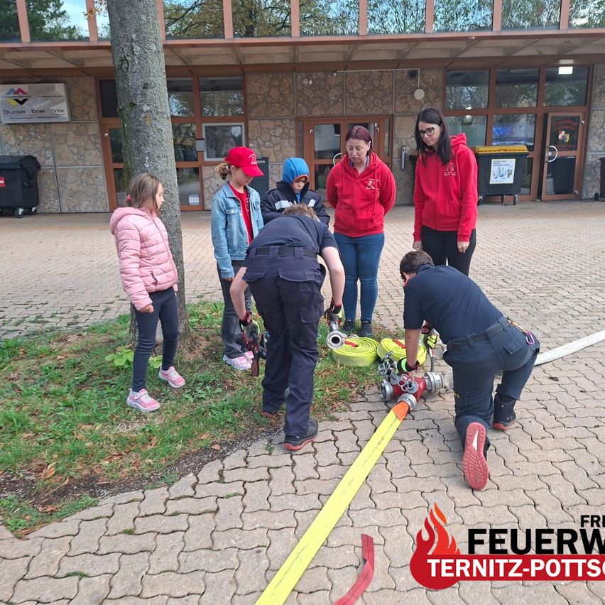 A group of children and adults are gathered around a fire hydrant, with two adults demonstrating its use. The children are wearing jackets and sneakers, and the adults are wearing red hoodies. The scene is outdoors, with a building in the background.