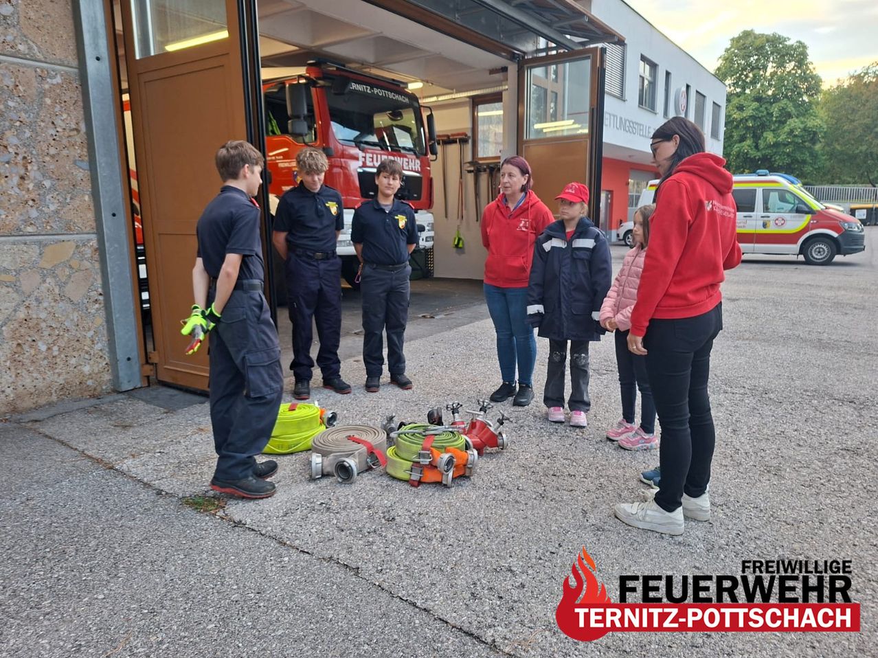 A group of firefighters and children stand outside a fire station, with fire hoses and equipment on the ground.