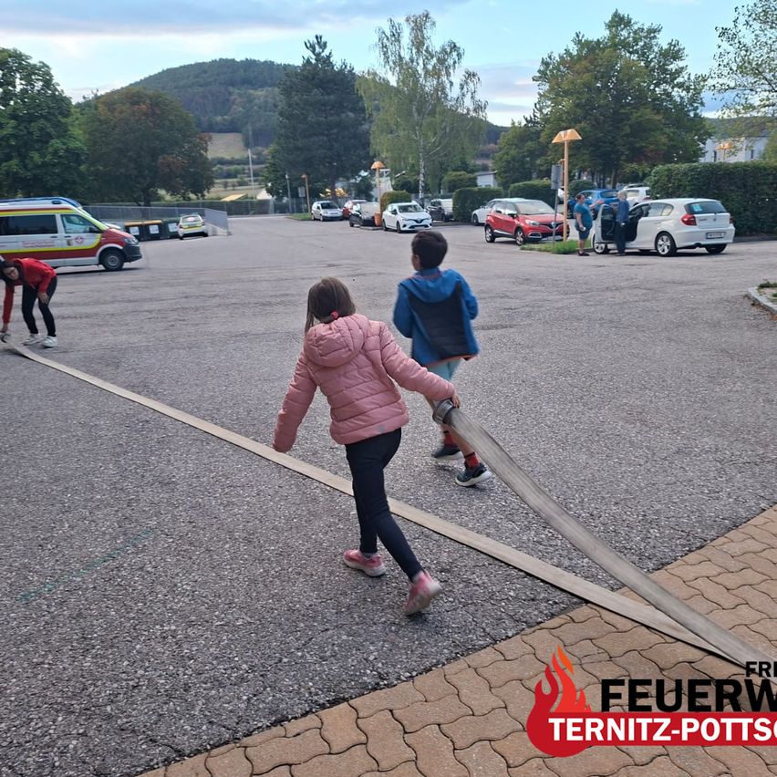 Children practice fire safety by pulling a fire hose in a parking lot with cars and trees in the background.