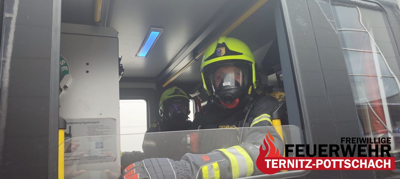 Two firefighters in protective gear inside a vehicle, one wearing a yellow helmet, the other with a green helmet, both equipped with masks.