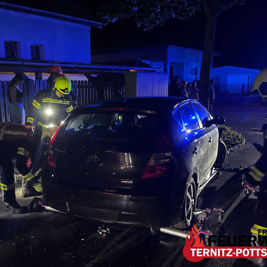 Firefighters work on a car accident at night in front of a house. A woman and several onlookers watch.