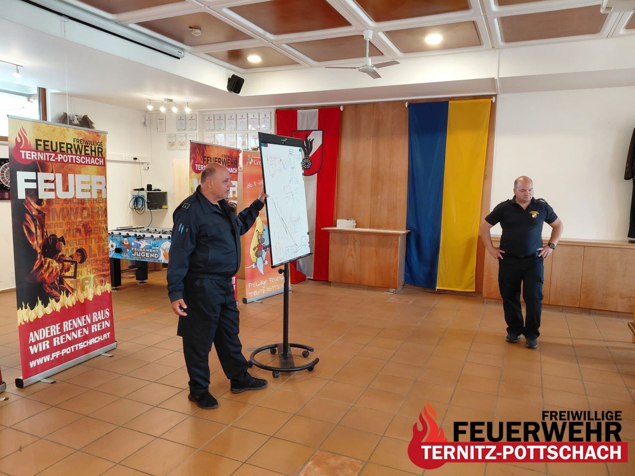 Two firefighters in uniform are in a training room. One points at a whiteboard. Flags and banners adorn the room, and the floor is tiled.