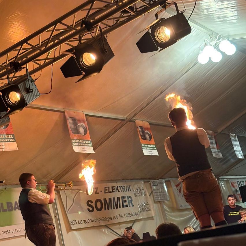 A man stands on a stage blowing a trumpet as fire ignites above him. Another man is in front of him, standing and blowing flames. Spotlights are overhead.