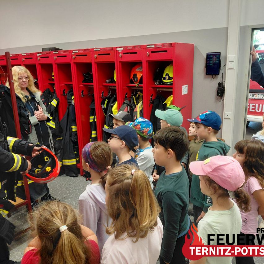 A firefighter holds a helmet while a group of children wearing hats and jackets watches from behind. Red lockers with yellow helmets hang behind them. A digital board is on the wall.
