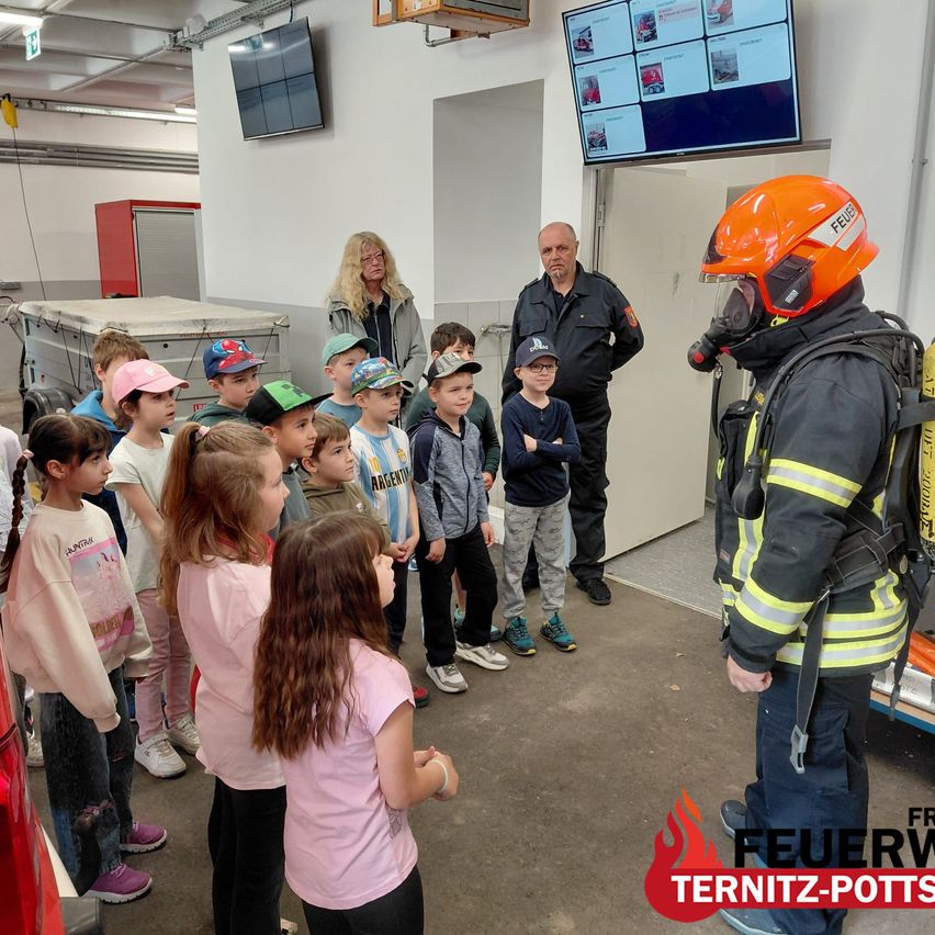 A firefighter in full gear talks to a group of children inside a fire station. Some adults stand behind the children.