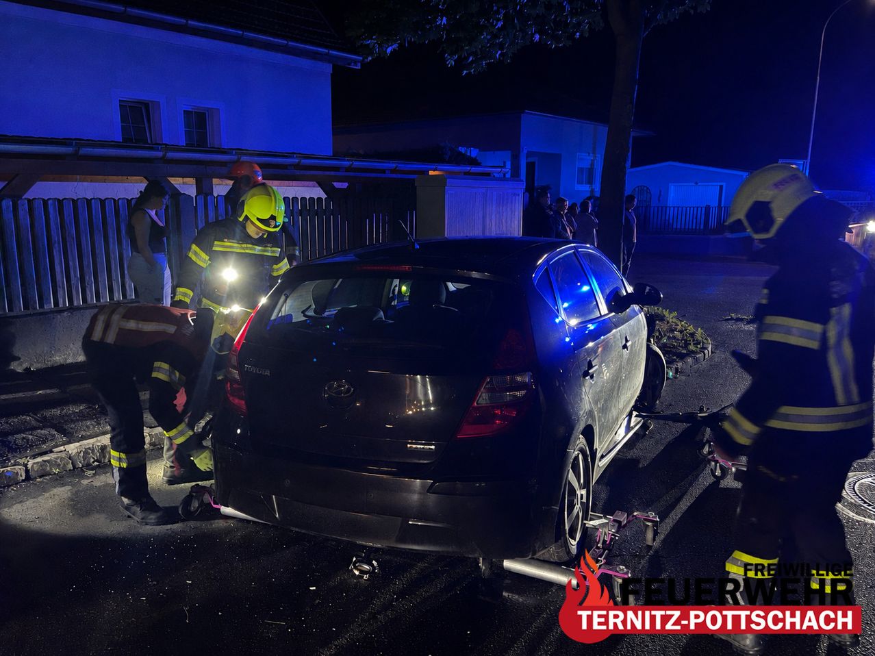 Firefighters are removing a person from a wrecked car on the street. They are using a car jack and have their lights on.