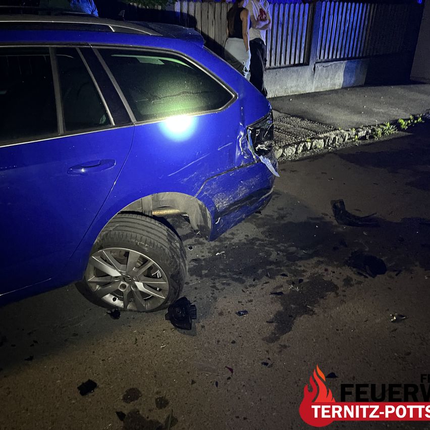 A blue car is damaged on the front side with some debris on the ground. There are two people standing behind the car.