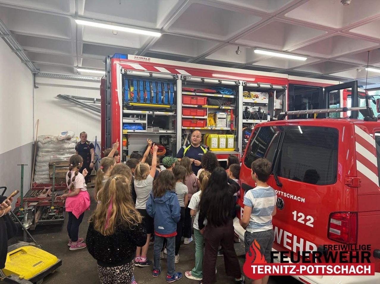 A group of children are gathered around a fire truck with a firefighter explaining something to them inside a fire station.