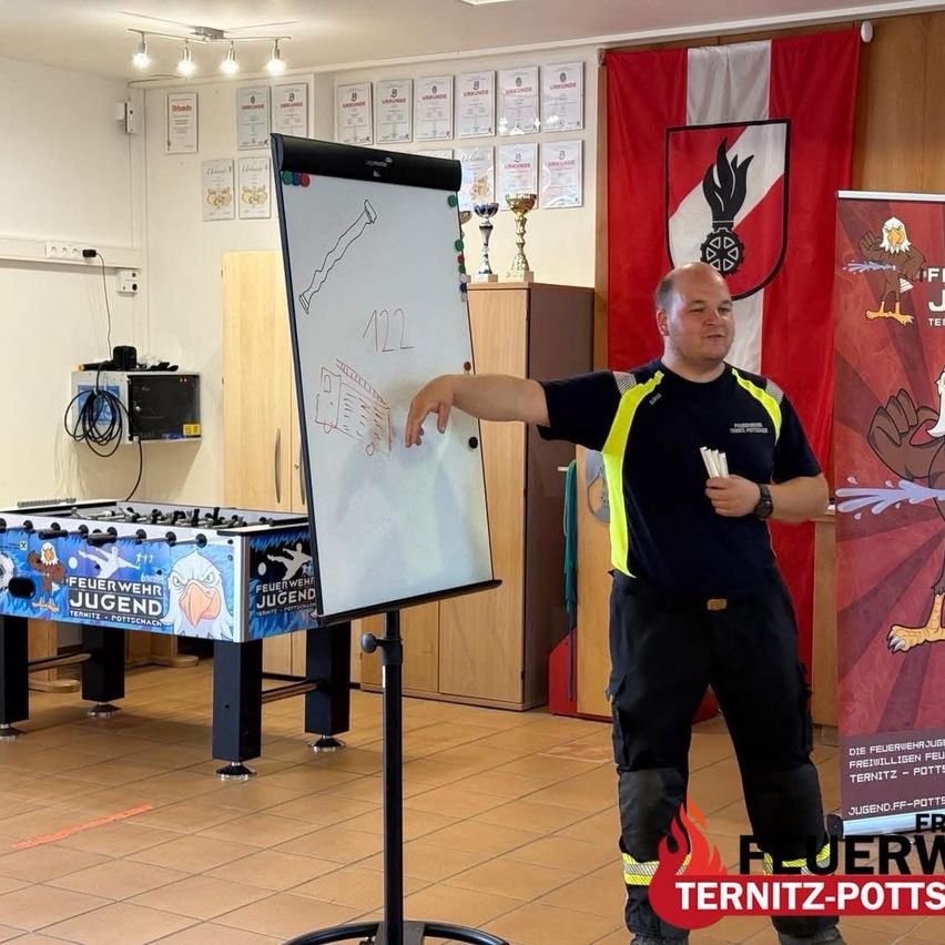 A man points to a whiteboard with a drawing and numbers while wearing a yellow and black uniform. A foosball table and a flag are in the background.