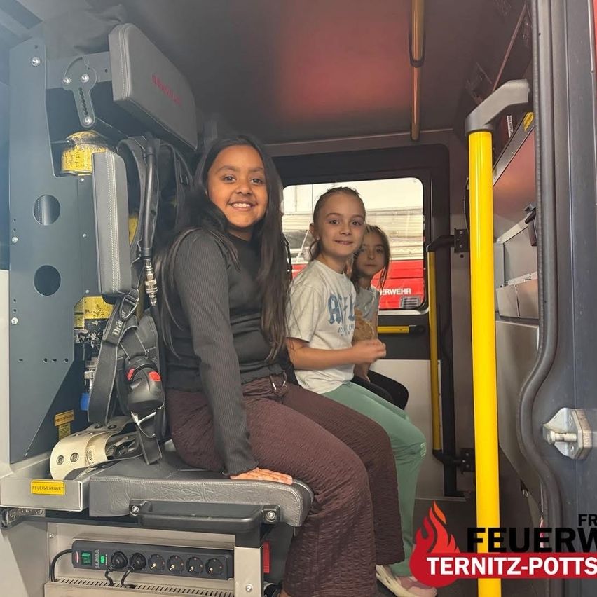 Three young girls sit inside a fire truck. One girl smiles for the camera, while the other two look out the window. Fire equipment and a yellow pole are visible.