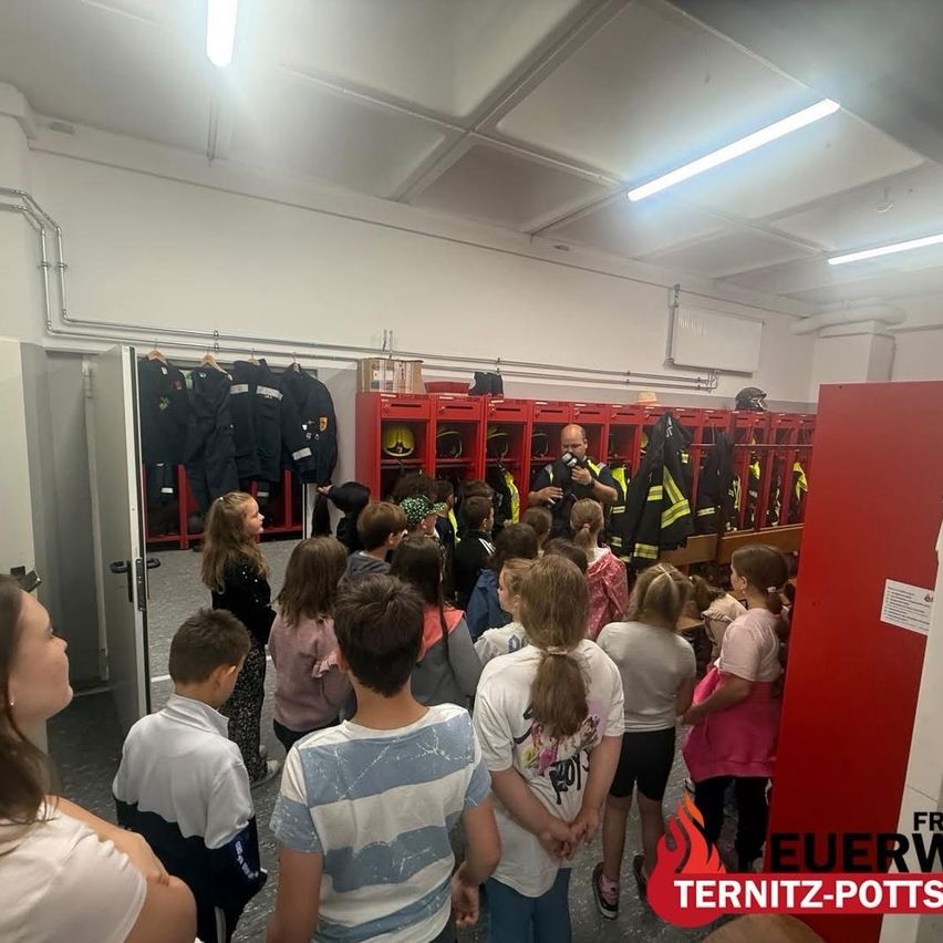 A group of children and adults stand inside a fire station, listening to a firefighter. Firefighting equipment is visible in the background.