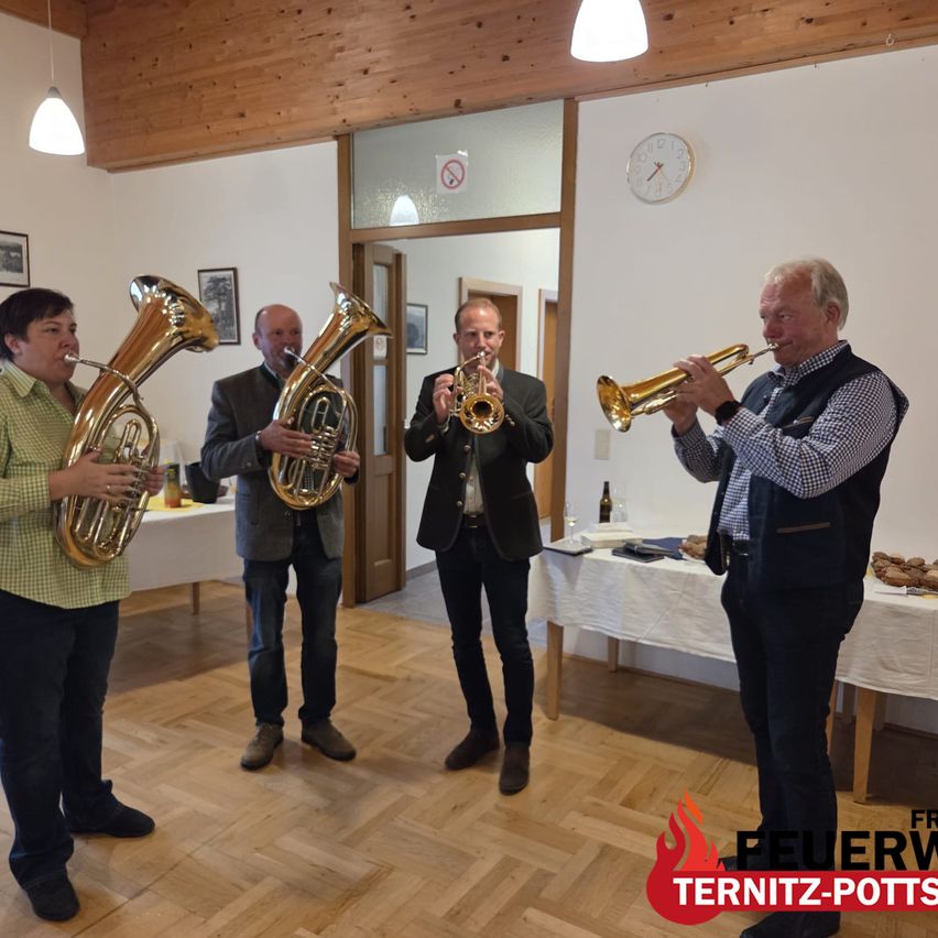 Vier Männer stehen in einem Raum und spielen Trompeten und Tubas. Ein Tisch mit Essen steht hinter ihnen, und eine Uhr ist an der Wand.