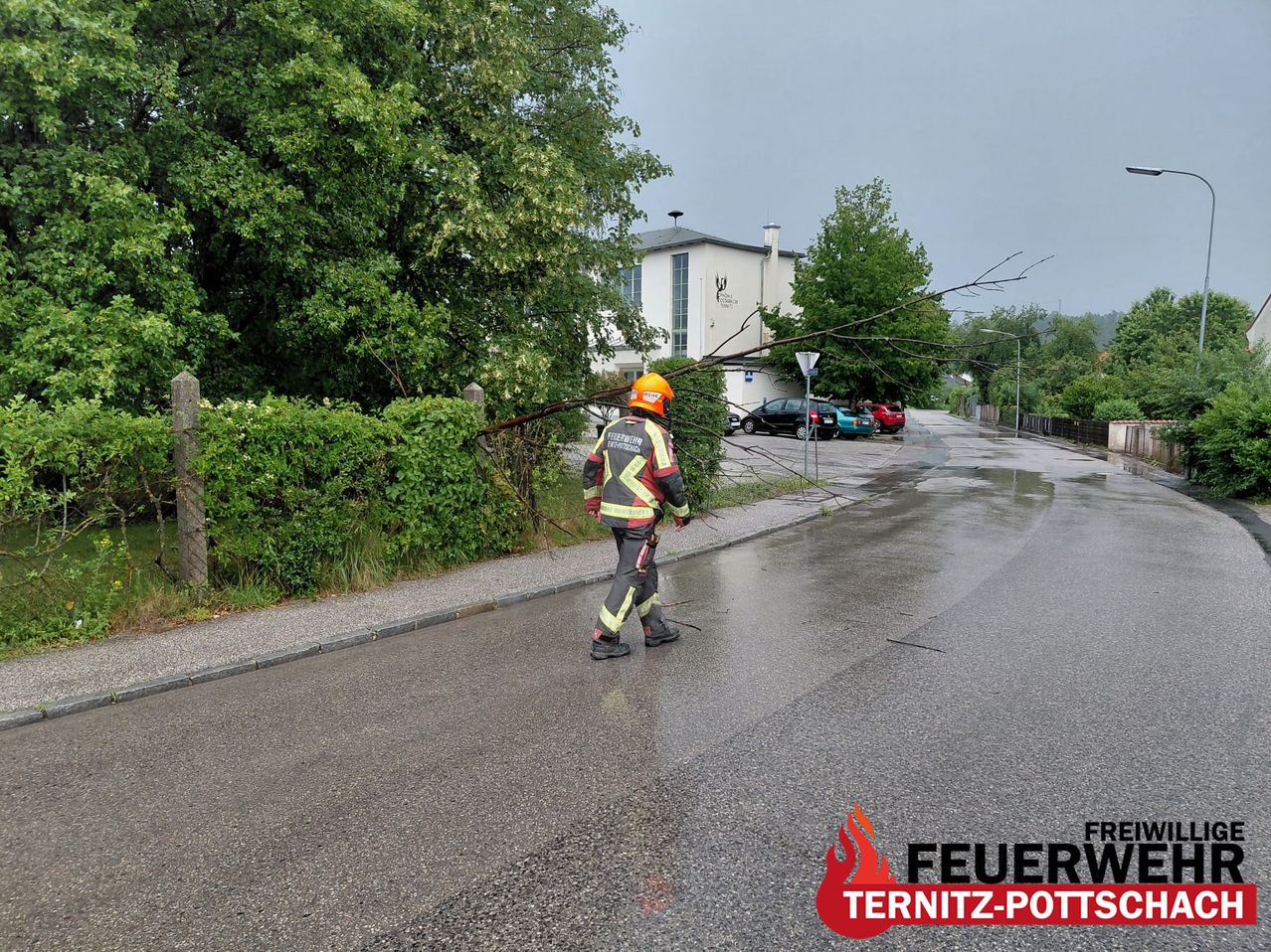 Ein Feuerwehrmann in voller Ausrüstung räumt Äste von einer nassen Straße. Dahinter sind ein weißes Gebäude und geparkte Autos zu sehen.