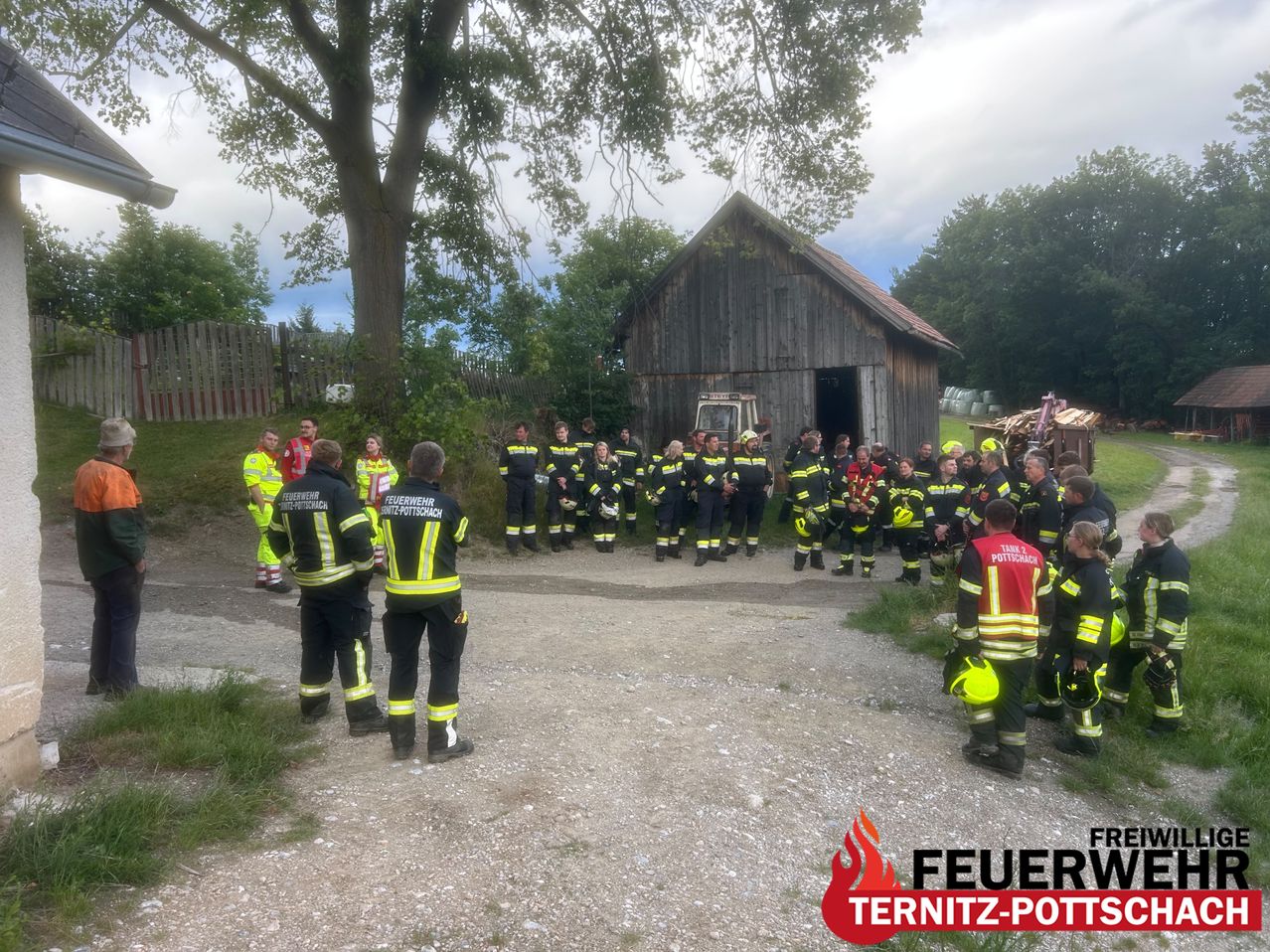 Eine Gruppe Feuerwehrleute in Uniform versammelt sich vor einer Scheune. Einige schauen in die Kamera, andere zur Scheune. Sie stehen auf einem Kiesweg.