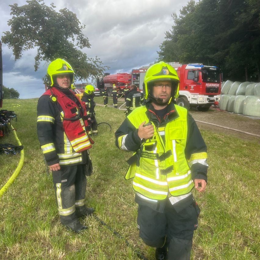 Zwei Feuerwehrleute in Schutzausrüstung stehen in einem Grasfeld, mit einem Feuerwehrauto und Heuballen im Hintergrund.