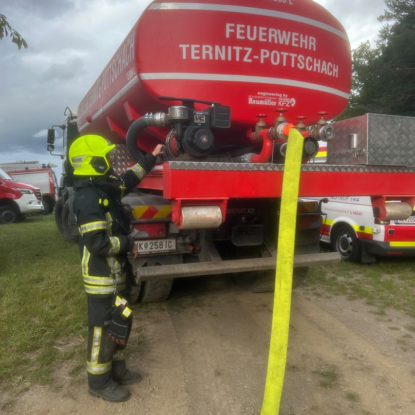 Ein Feuerwehrmann in voller Montur steht neben einem roten Feuerwehrwagen mit den Worten 'Feuerwehr Ternitz-Pottschach' darauf. Er hält einen gelben Schlauch in der Nähe des Fahrzeugs.